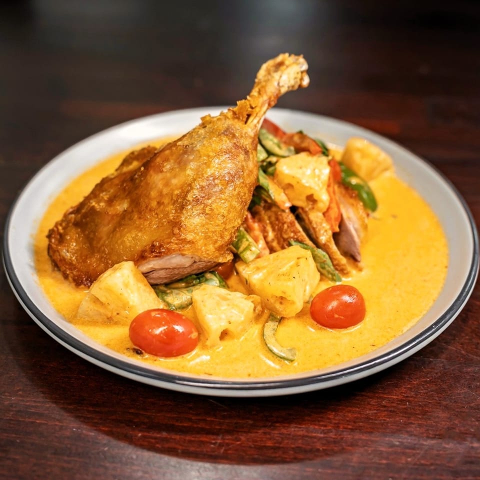 Crispy duck leg in red curry with pineapple, tomatoes, and bell peppers on a white plate.