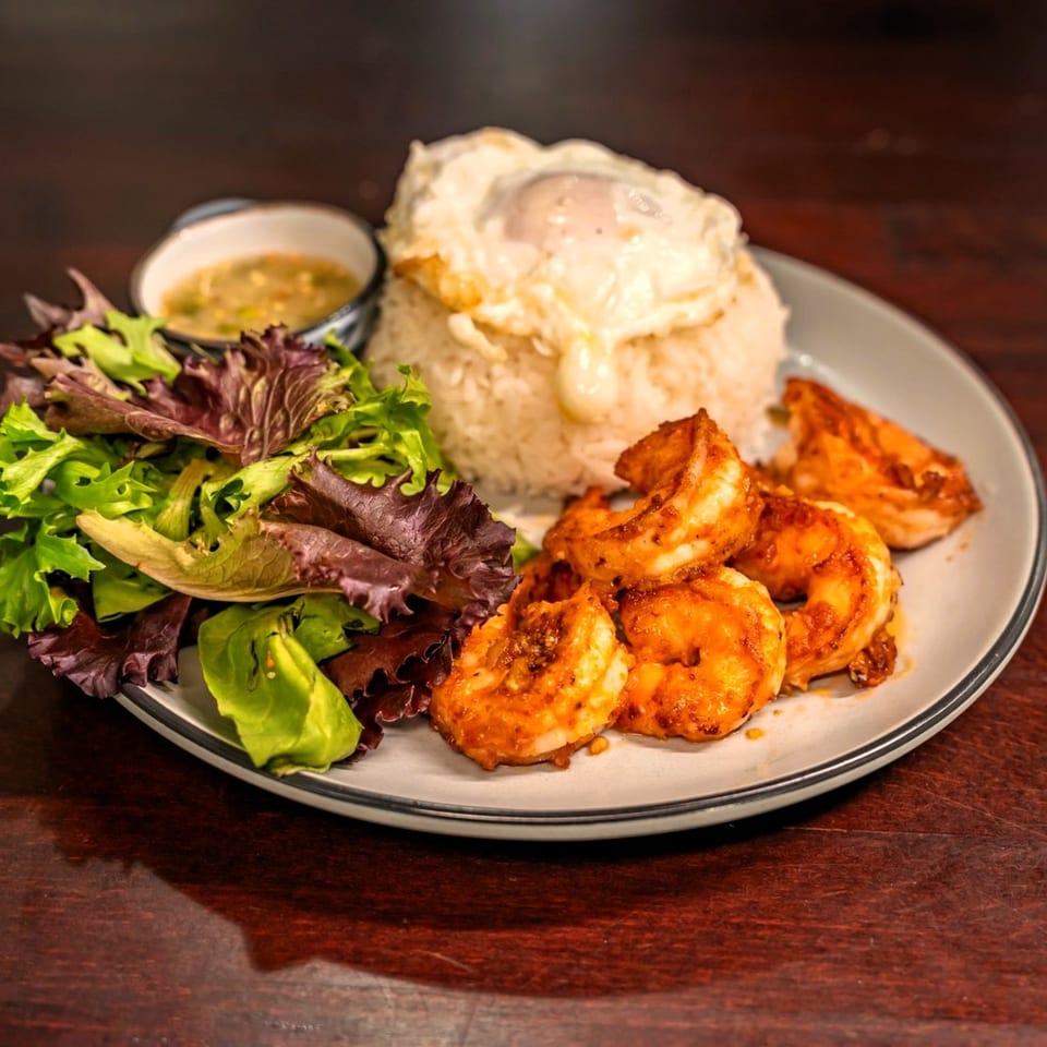 Thai dish with rice, fried egg, seasoned shrimp, fresh salad, and a side of sauce.